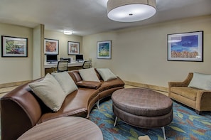 A hotel room with a brown leather sofa, a round ottoman, a small armchair, a desk with a computer, and framed artwork on the walls. - Candlewood Suites Louisville Airport by IHG (Louisville)