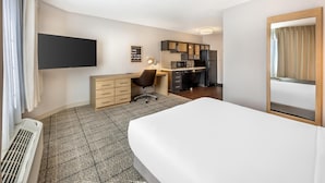 Habitación de hotel con cama, escritorio, kitchenette y televisor montado en la pared.