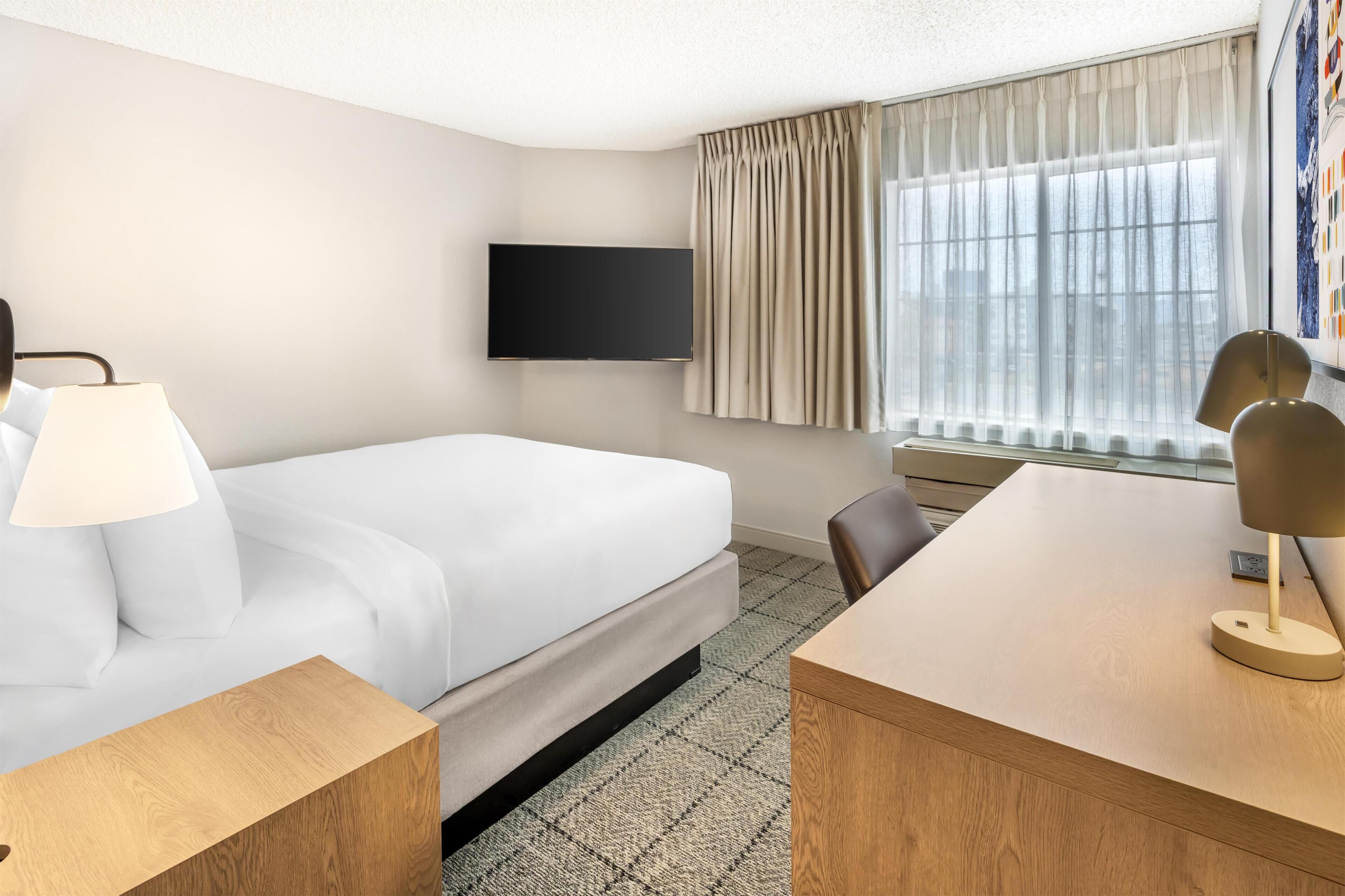 A hotel room with a bed, a desk, a chair, a TV mounted on the wall, and a window with curtains.