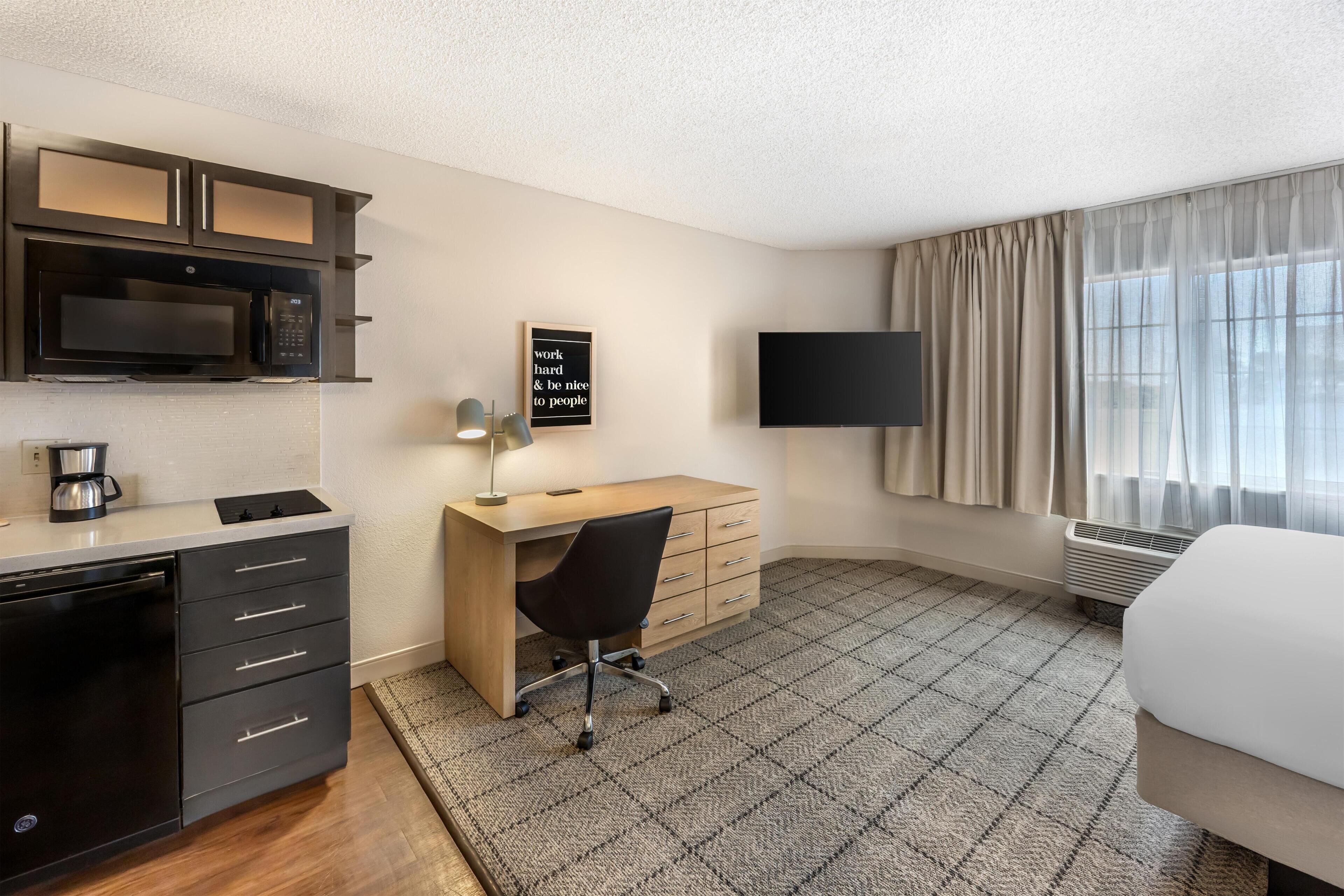 Une chambre d’hôtel équipée d’une kitchenette, d’un bureau, d’une chaise, d’un lit, d’une télévision et d’une fenêtre avec des rideaux.