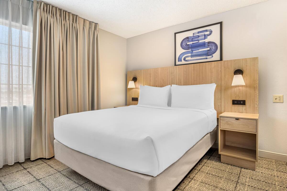 Een hotelkamer met een groot bed, twee bedlampen, een houten hoofdeinde, een kunstwerk in een lijst en een vloerbedekking.
