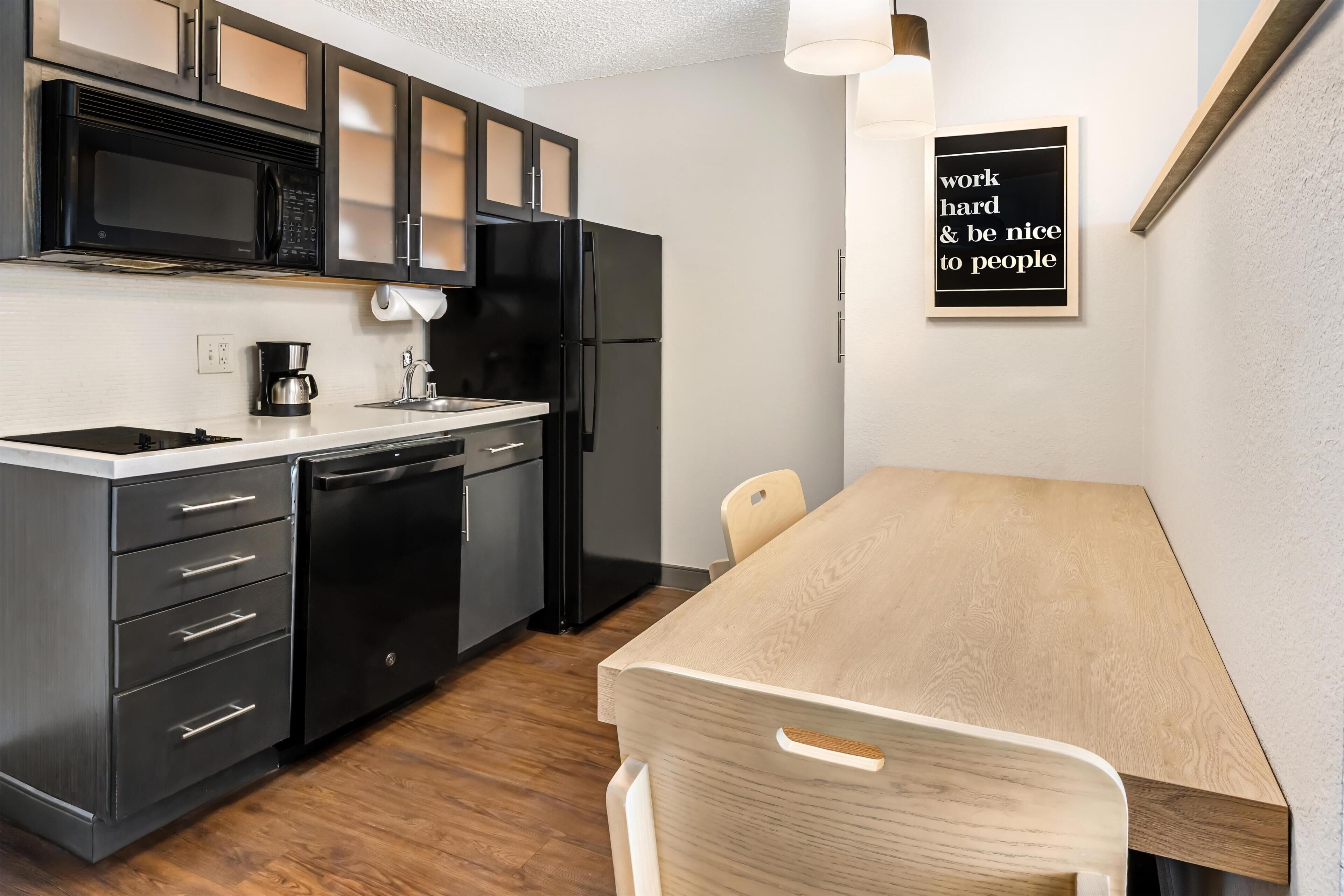 A modern kitchen with black appliances, a wooden dining table, and a motivational quote on the wall.