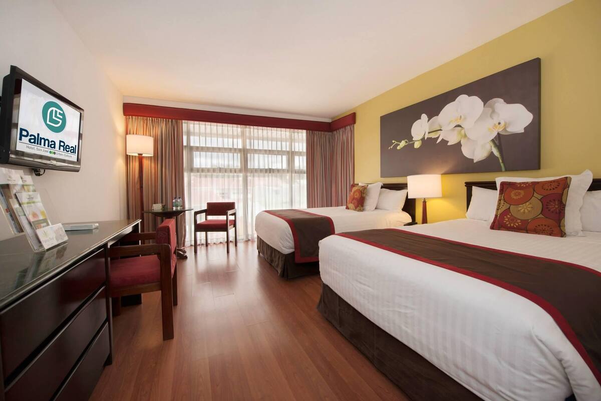 A hotel room with a desk, two beds, a TV, and a large orchid painting.