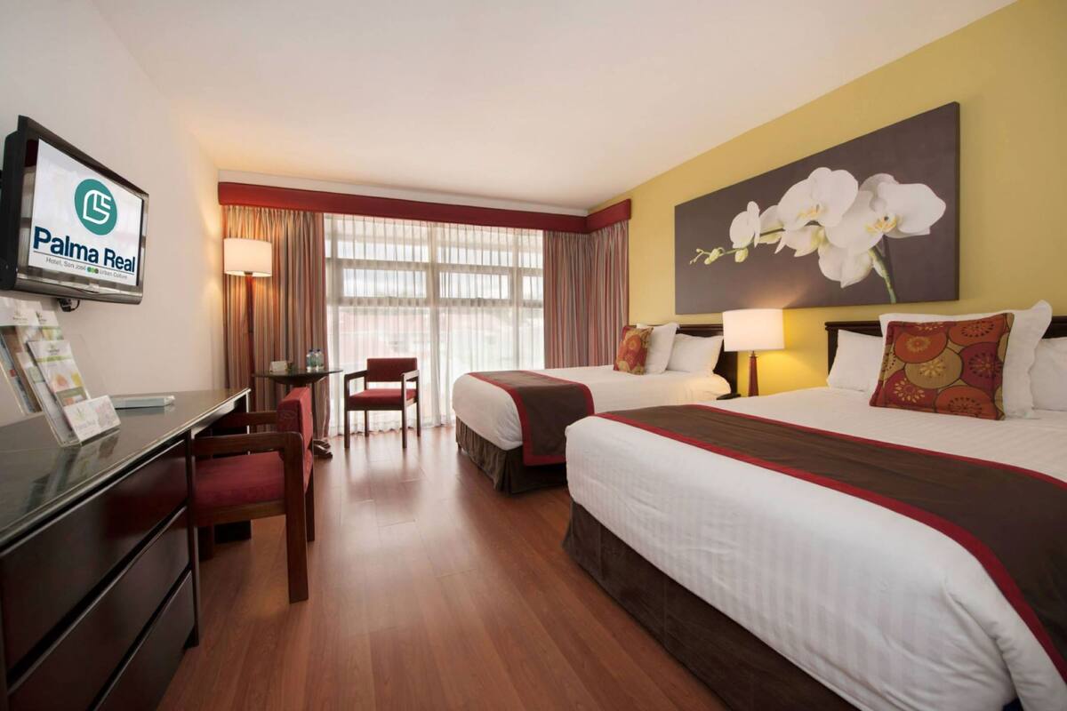 Habitación de hotel con dos camas, un escritorio, una silla, un televisor y un cuadro grande de una orquídea en la pared.