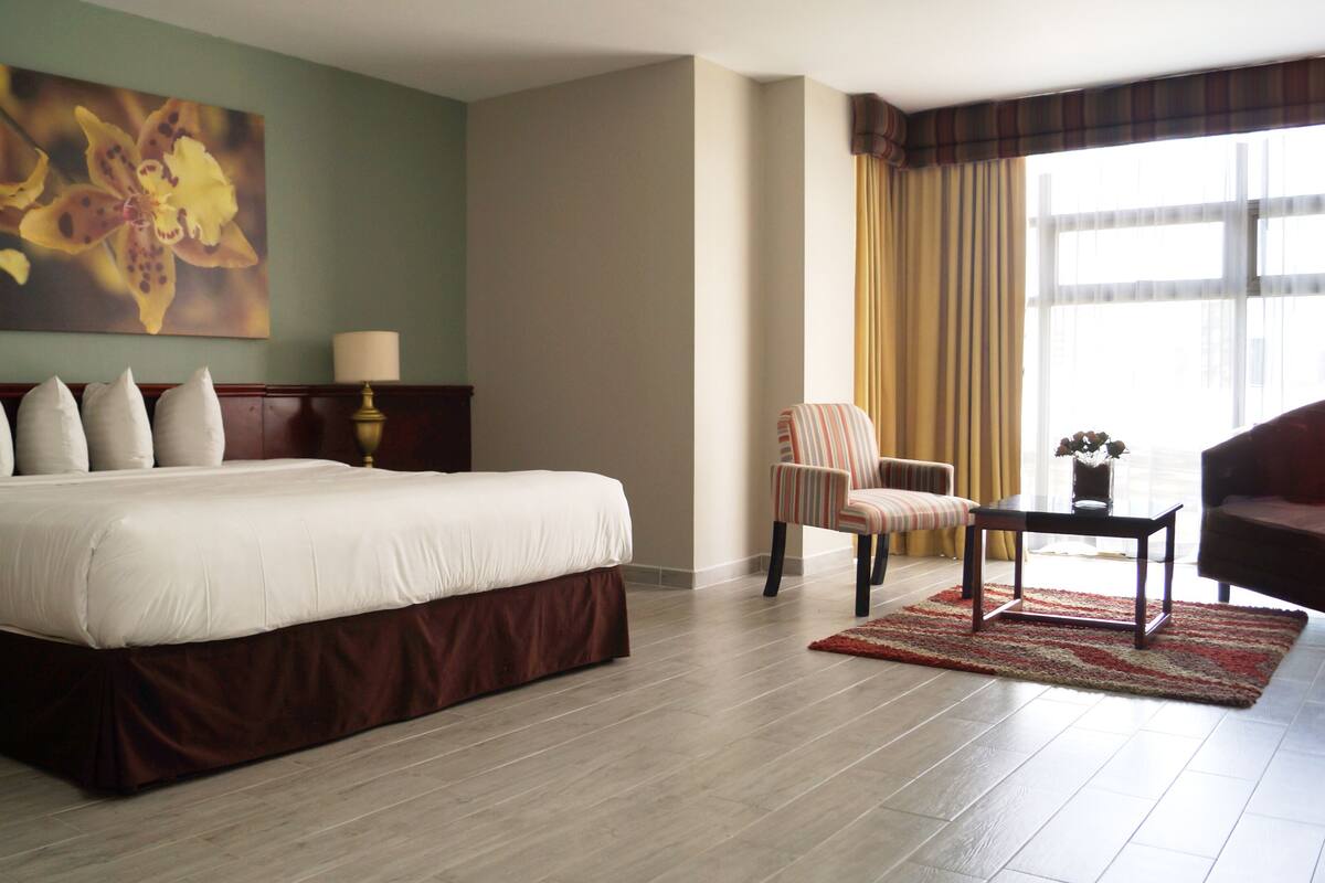 Habitación de hotel con una cama grande, una silla, una mesita con un florero y un cuadro en la pared.