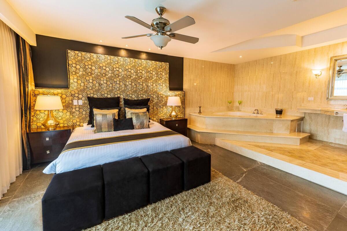 A modern hotel room with a large bed, a sitting area, a bathtub, and a bathroom with a vanity and mirror.