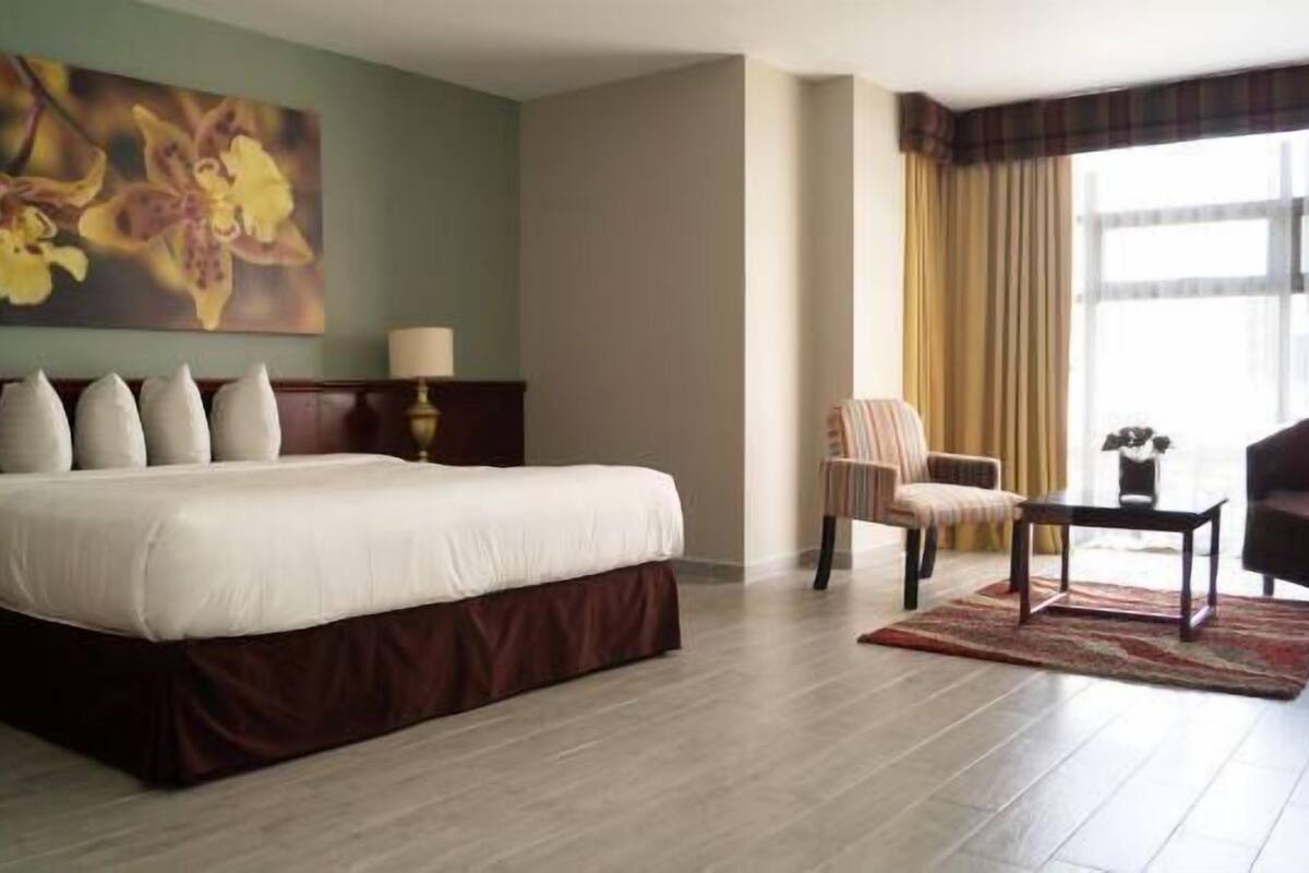 A hotel room with a large bed, a chair, a small table, and a painting on the wall.
