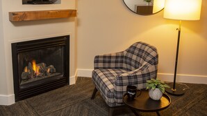A living room with a fireplace, a plaid armchair, a small table with a plant and a mug, a floor lamp, and a mounted TV.
