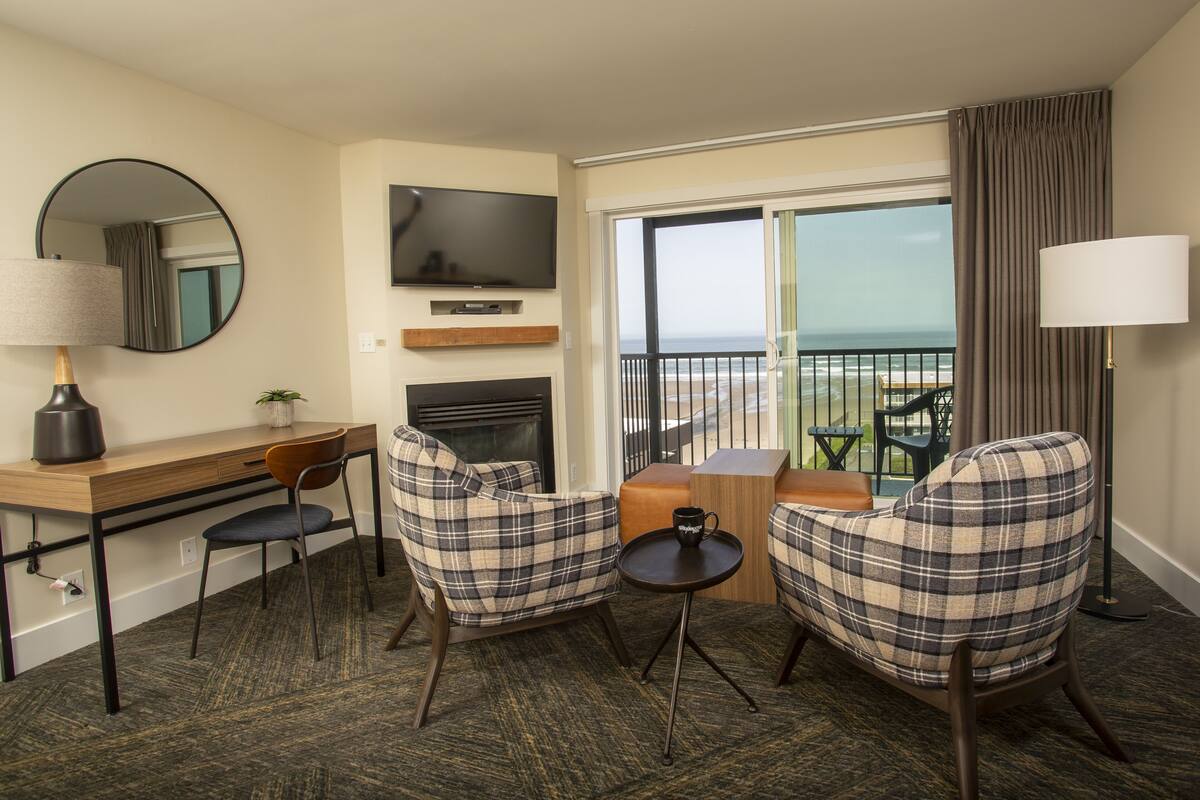 Hotelkamer met een bureau, twee geruite fauteuils, een tafeltje met een mok, een televisie en een balkon met uitzicht op de oceaan.