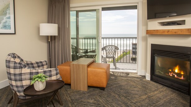 A hotel room with a sofa, a small table with a plant, a fireplace, a television, and a balcony with a view of the ocean.