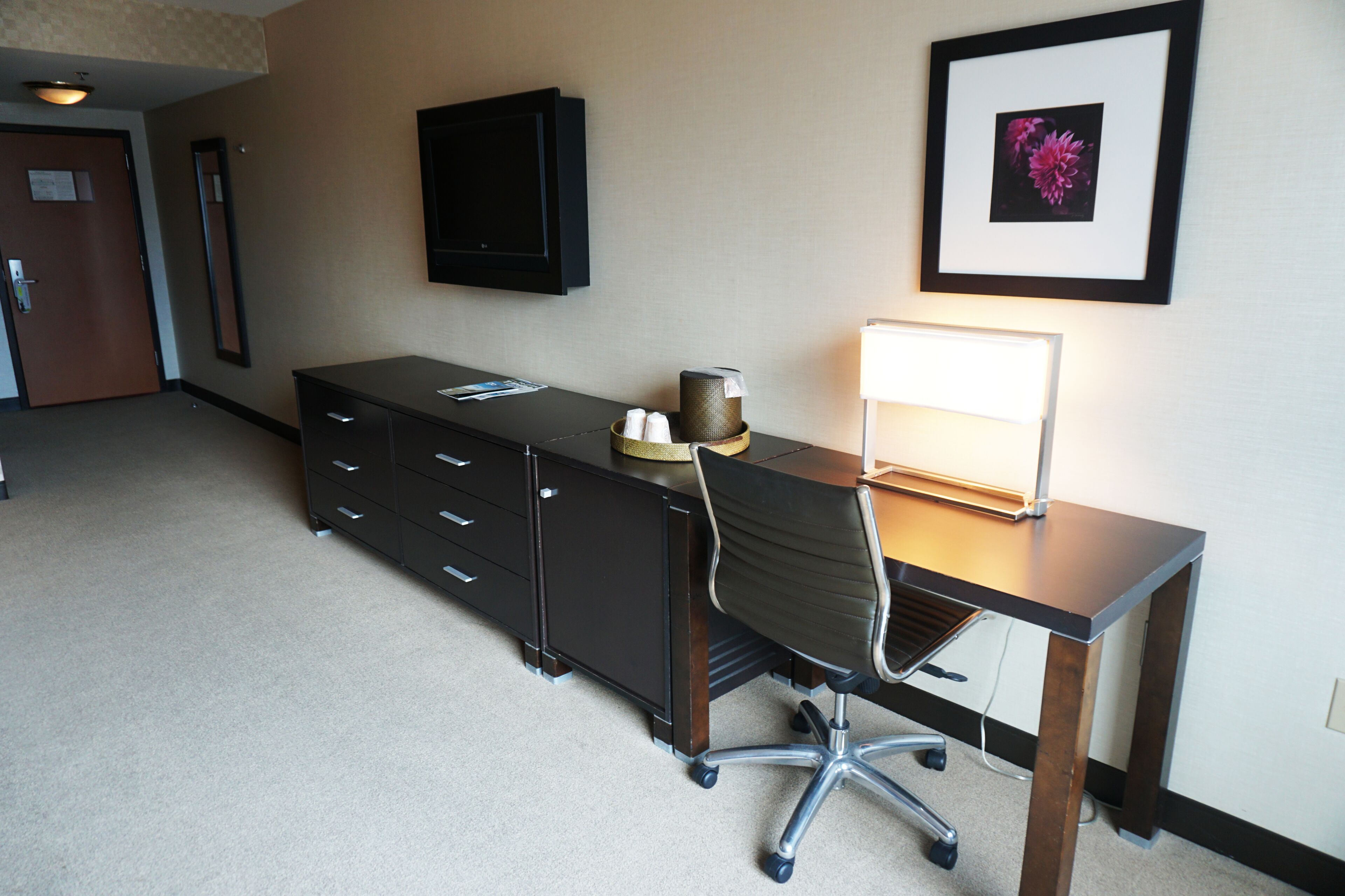 A hotel room with a desk, chair, and a flat-screen TV mounted on the wall.
