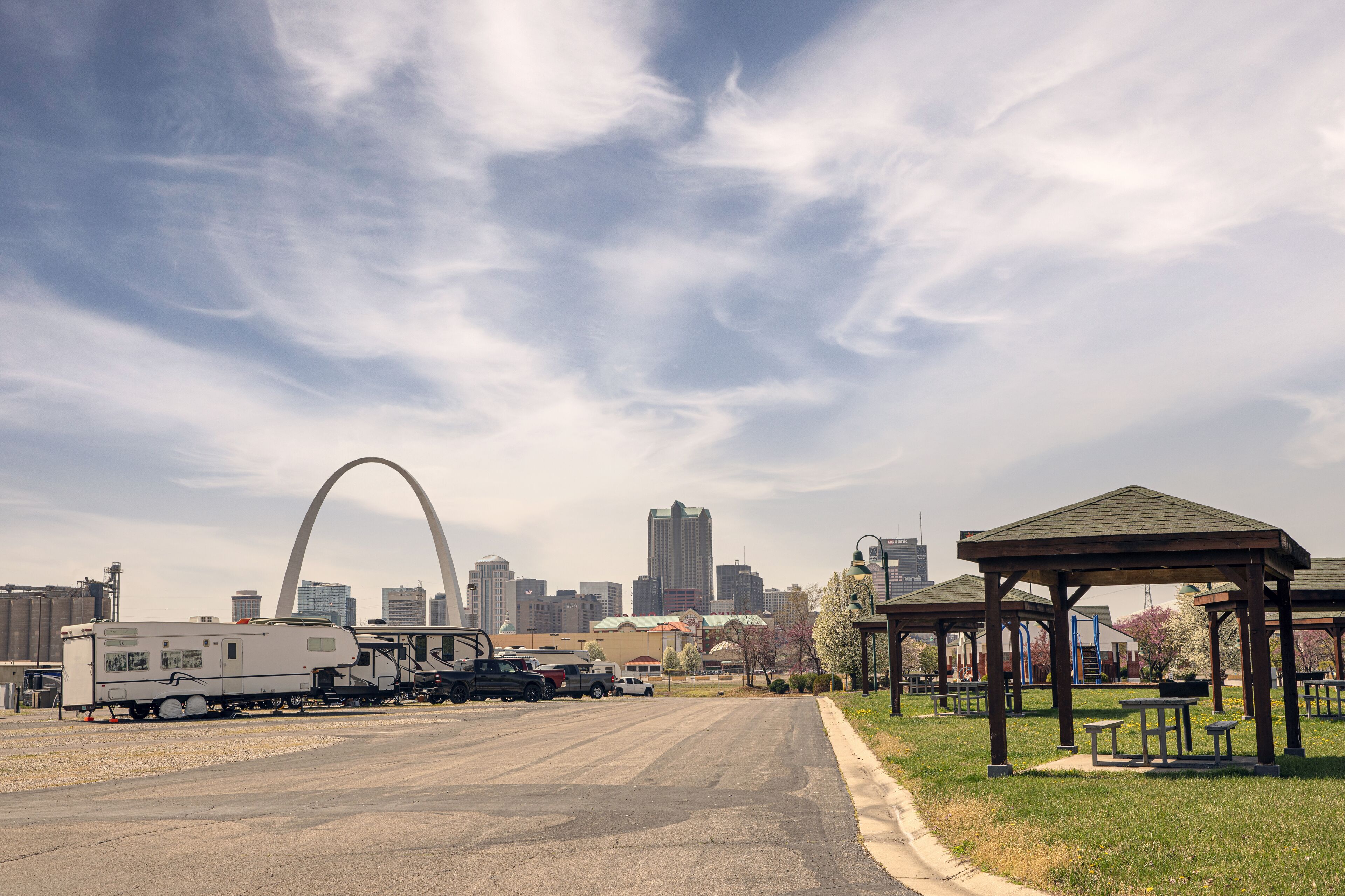 A cityscape with a prominent arch structure, a park with a gazebo, and a train with RVs parked nearby.