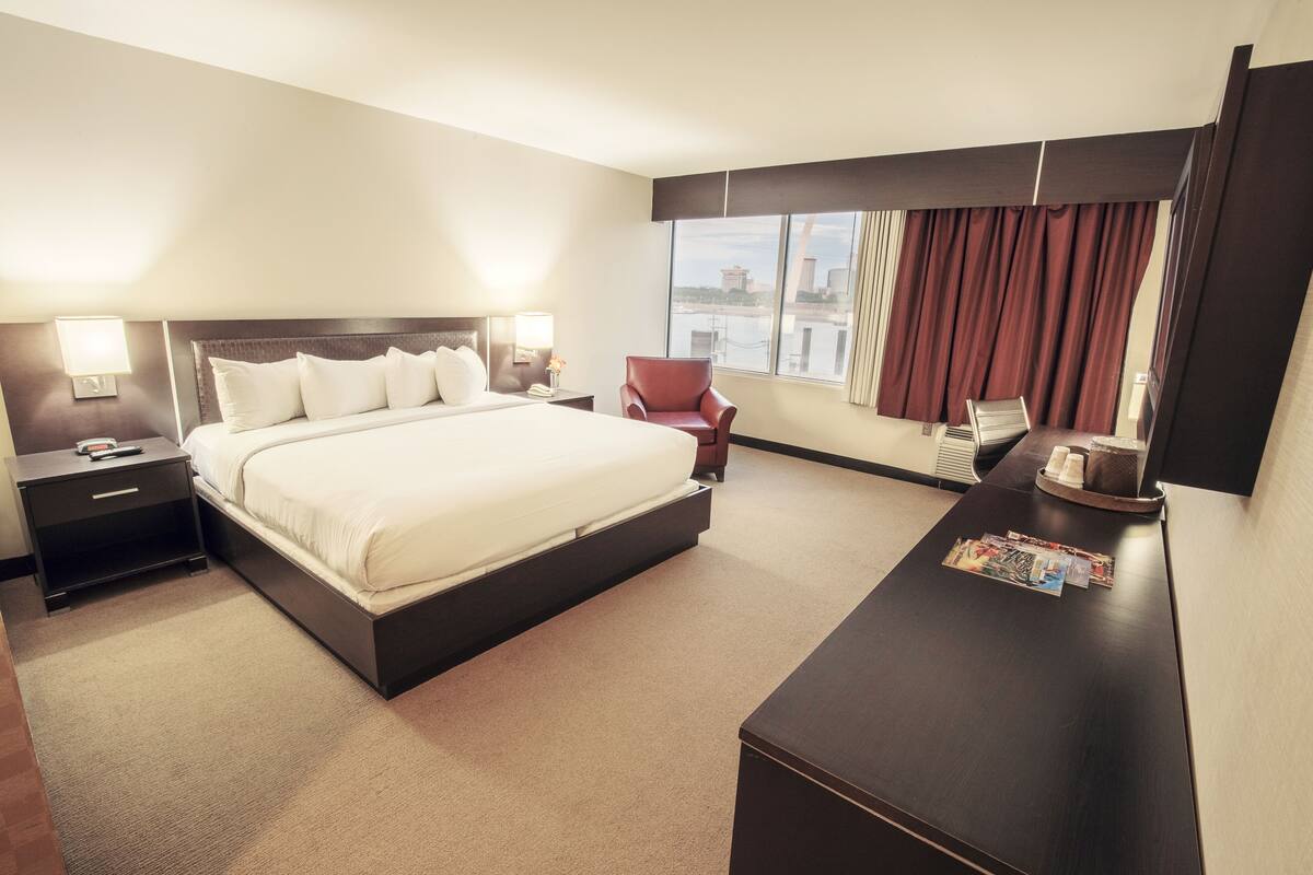 A hotel room with a large bed, a desk with a chair, a nightstand, and a window with curtains.