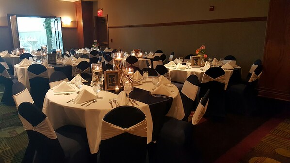 Un salón de banquetes con mesas redondas dispuestas para comer, cada una con mantel blanco, sillas negras y centros de mesa.