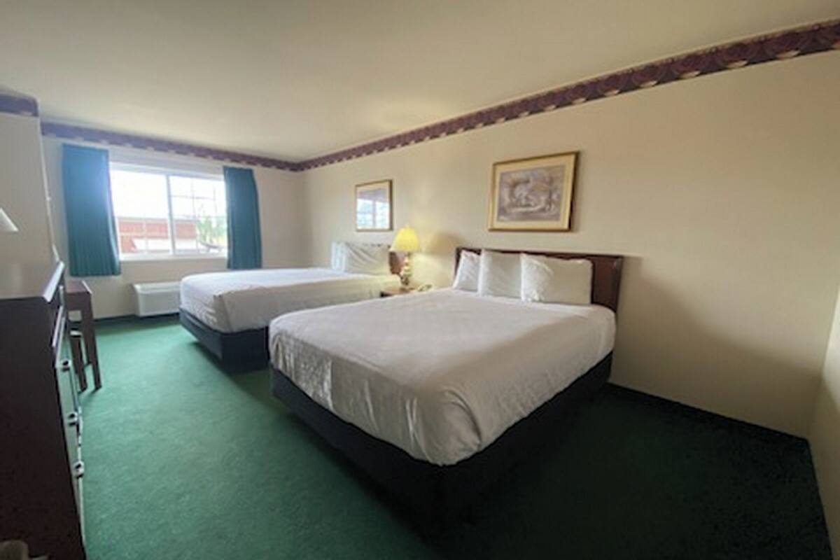 A hotel room with two beds, a desk, a chair, a window with curtains, and a framed picture on the wall.