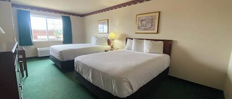 A hotel room with two beds, a desk, a chair, a window with curtains, and a framed picture on the wall.