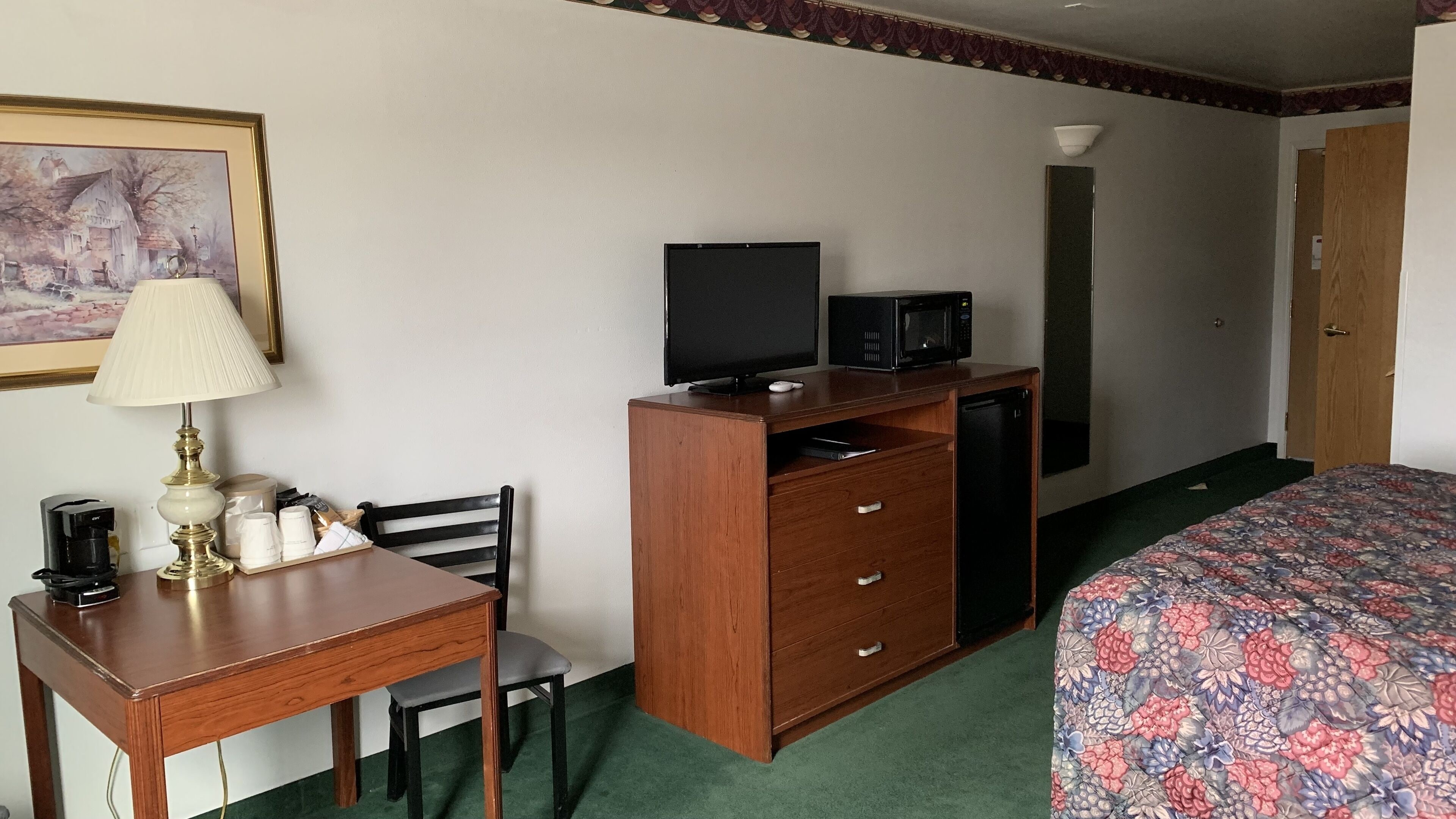 A hotel room with a wooden desk, a chair, a television, a microwave, a bed with a floral bedspread, and a painting on the wall.