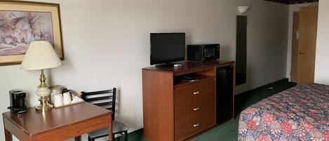 A hotel room with a wooden desk, a chair, a television, a microwave, a bed with a floral bedspread, and a painting on the wall.