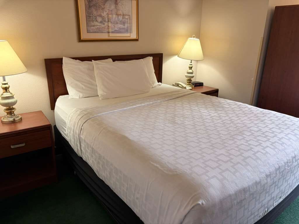 A neatly made bed with white linens, two bedside lamps, a wooden headboard, and a framed picture on the wall.