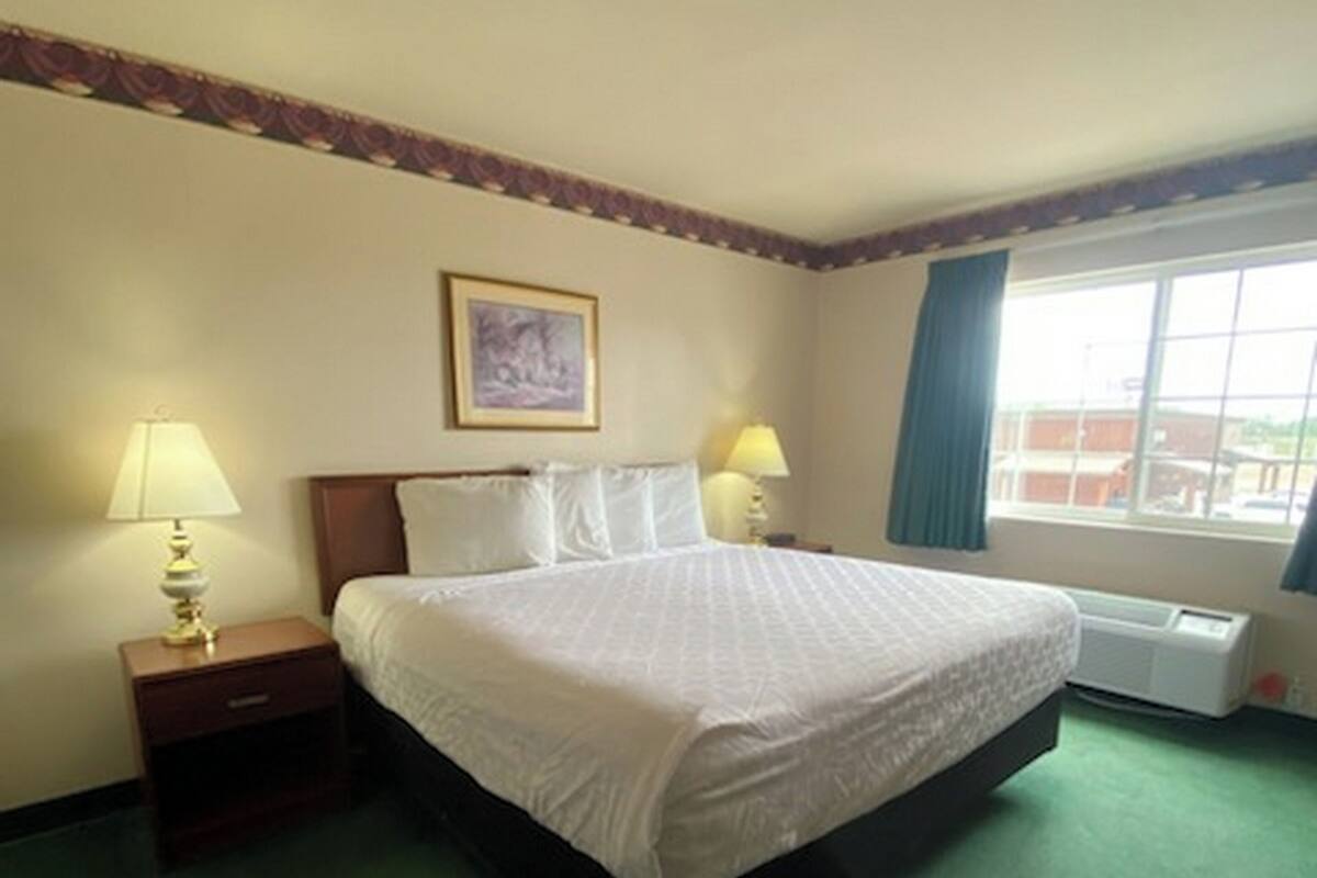 A hotel room with a large bed, two bedside tables with lamps, a window with curtains, and a framed picture on the wall.