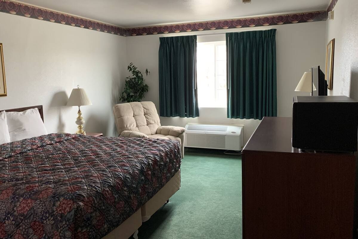 A hotel room with a large bed, a chair, a desk with a TV, an air conditioner, and a window with curtains.