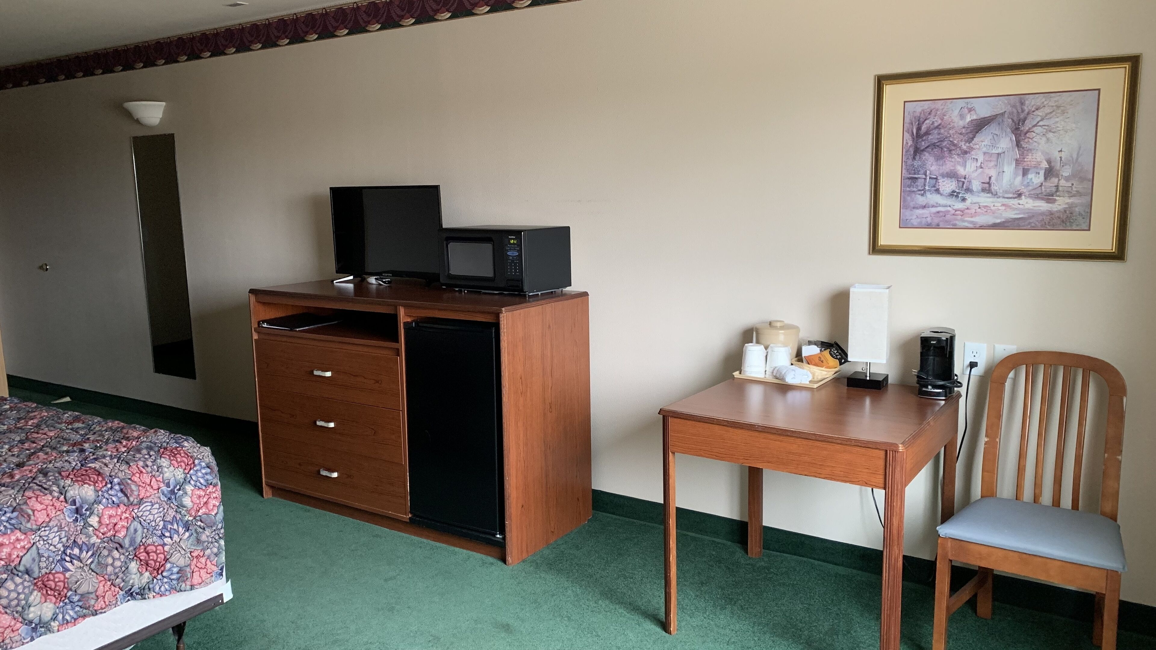 A hotel room with a bed, a desk with a chair, a microwave, a small refrigerator, and a painting on the wall.