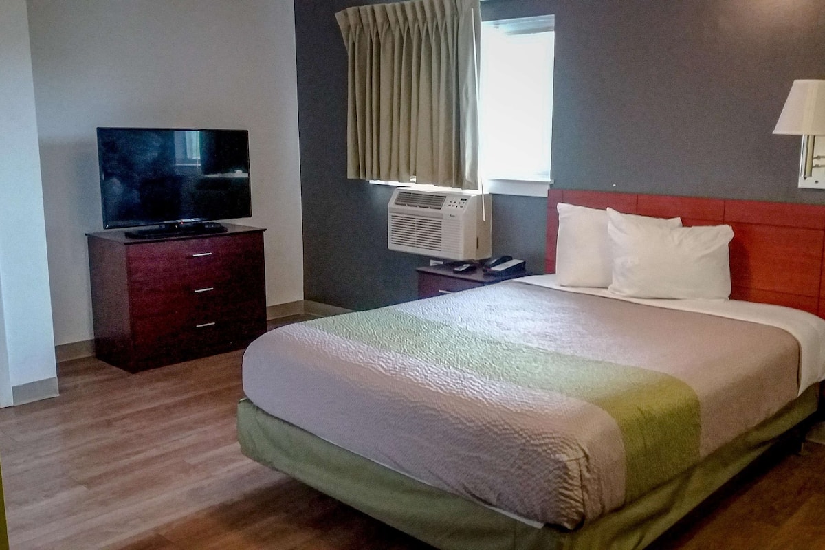 A hotel room with a bed, a TV on a wooden dresser, an air conditioning unit, a window with curtains, and a bedside lamp.