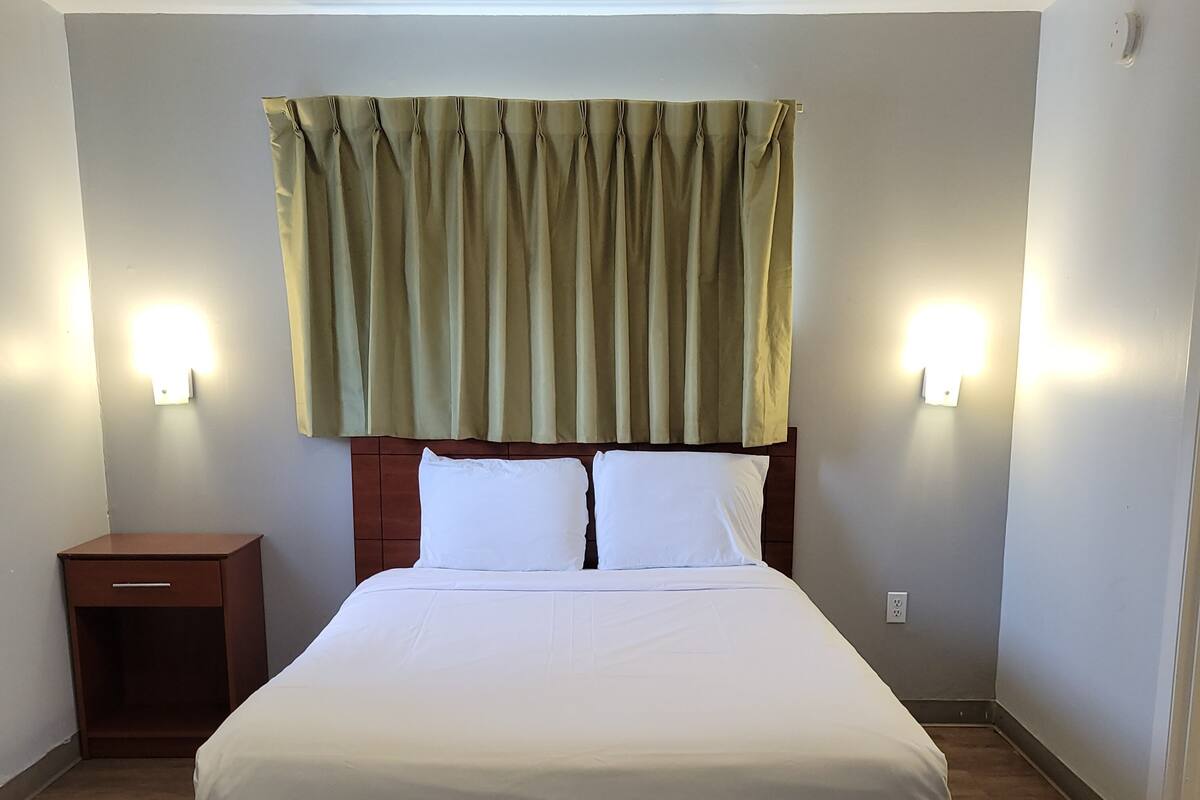 A large bed with white linens, a wooden headboard, and a green curtain. There are two wall-mounted lights on either side of the bed.