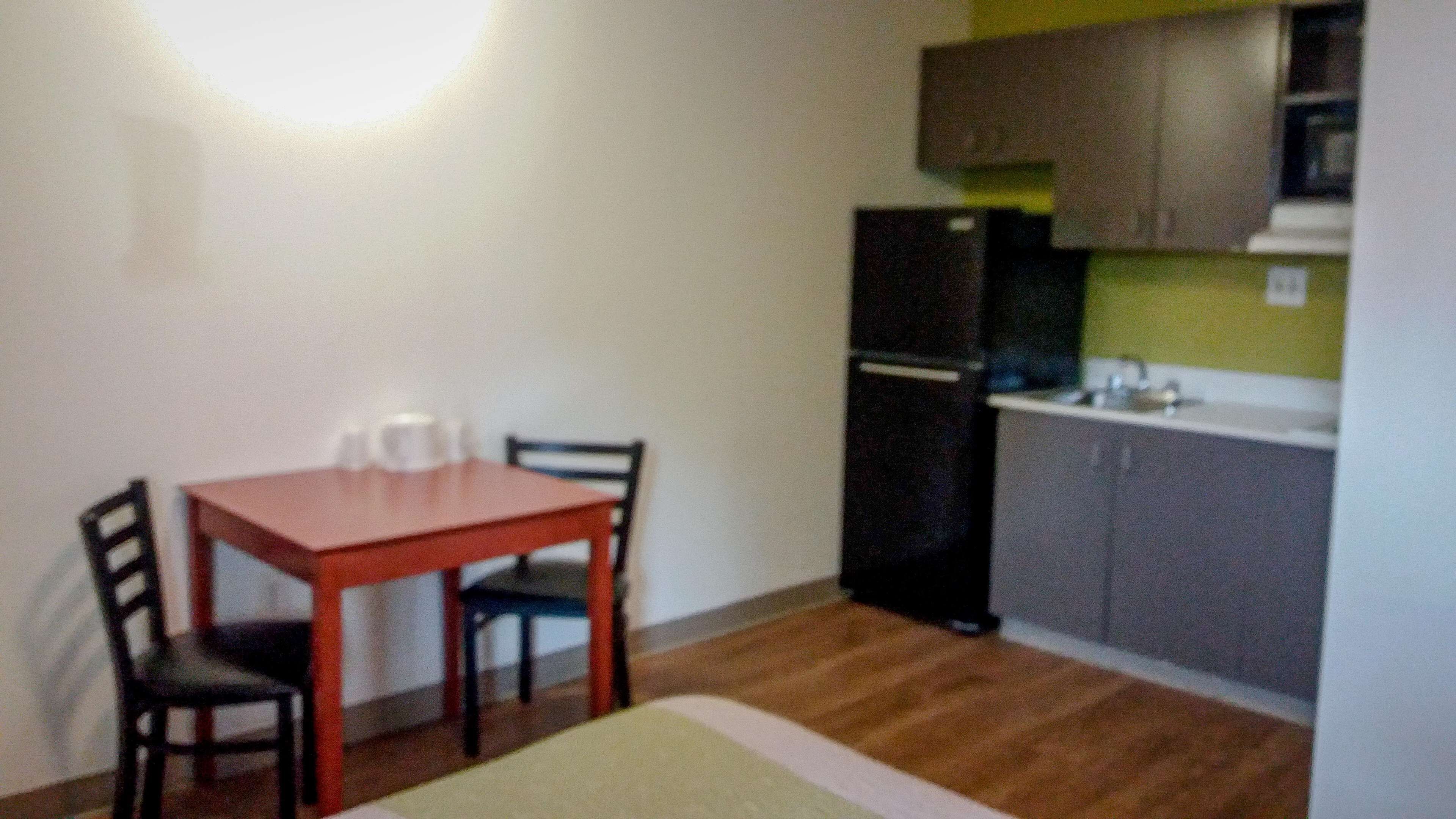 Una pequeña cocina americana equipada con refrigerador, lavamanos y alacenas. Hay una mesa de comedor con sillas y una alfombra en el suelo.