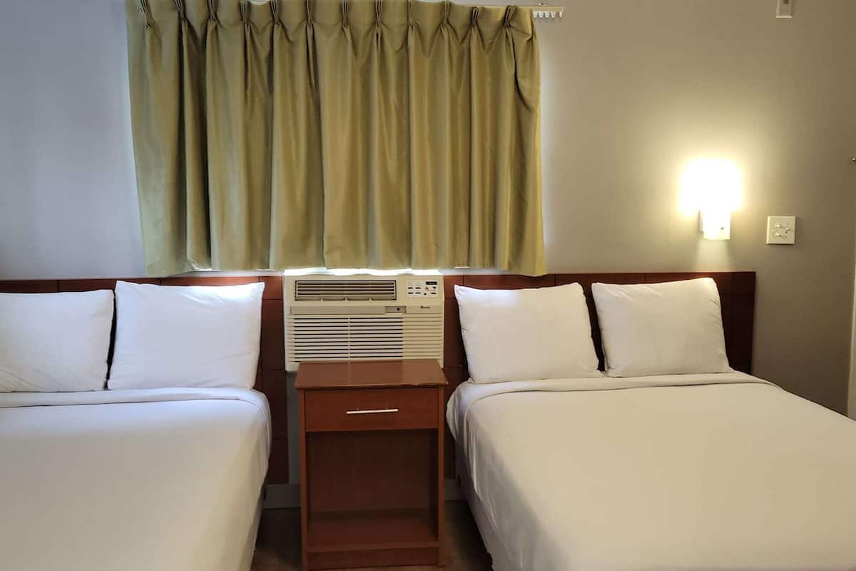 A hotel room with two single beds, a nightstand, an air conditioning unit, a window with curtains, and a wall-mounted light.