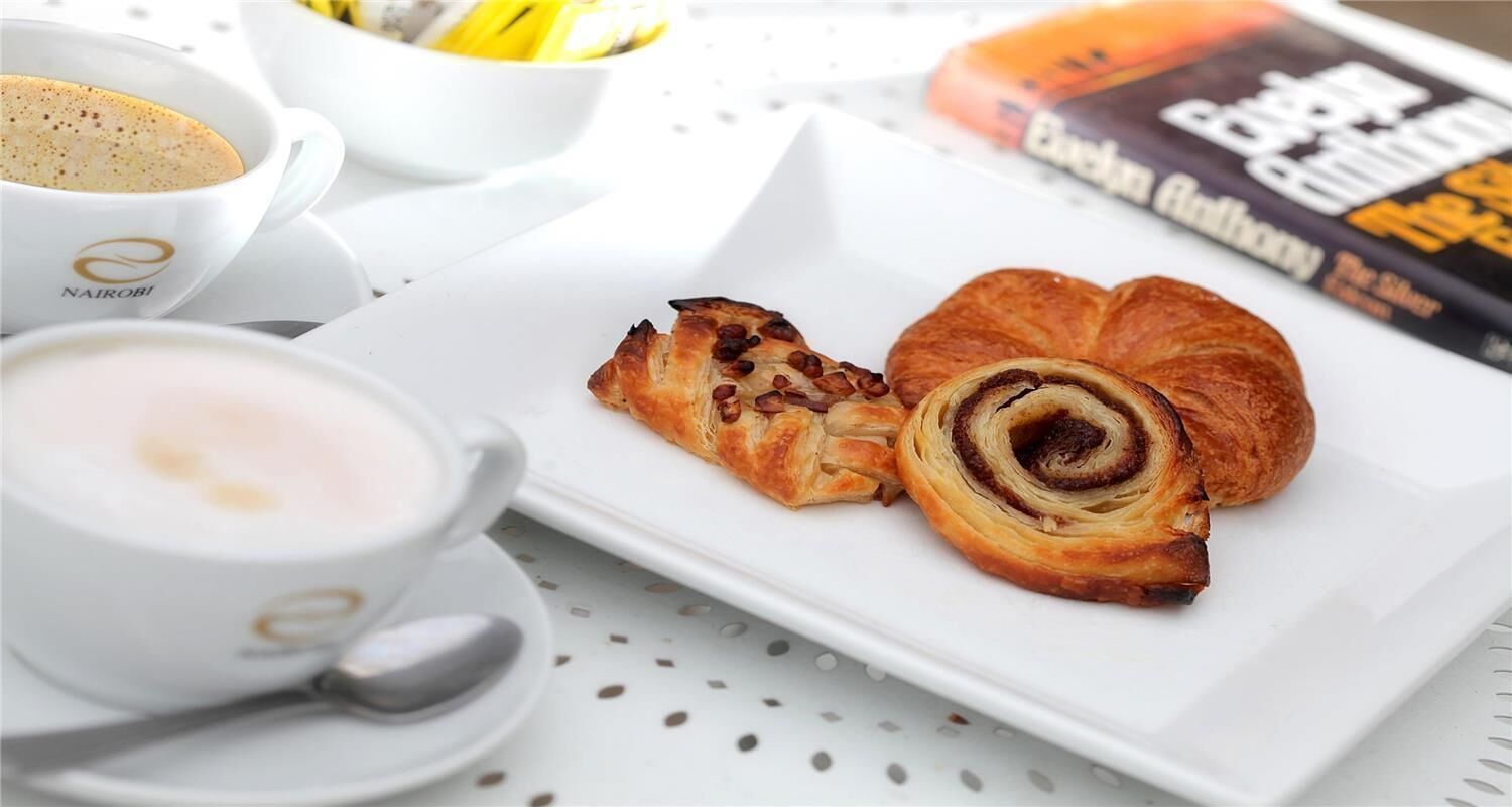 Café da manhã com um croissant e uma xícara de café em um prato branco, uma colher e uma revista.