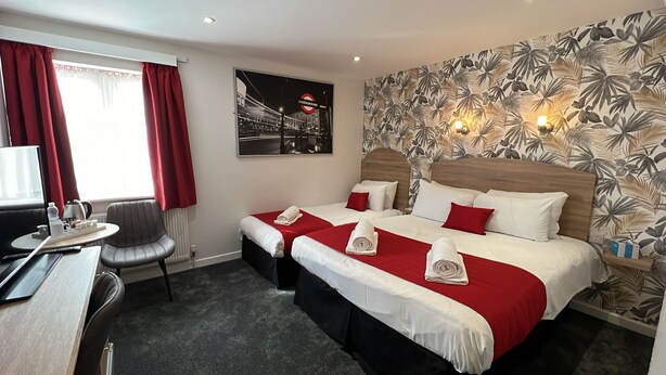 Ein Hotelzimmer mit zwei Betten, einem Schreibtisch, einem Stuhl und einem Fenster mit roten Vorhängen.
