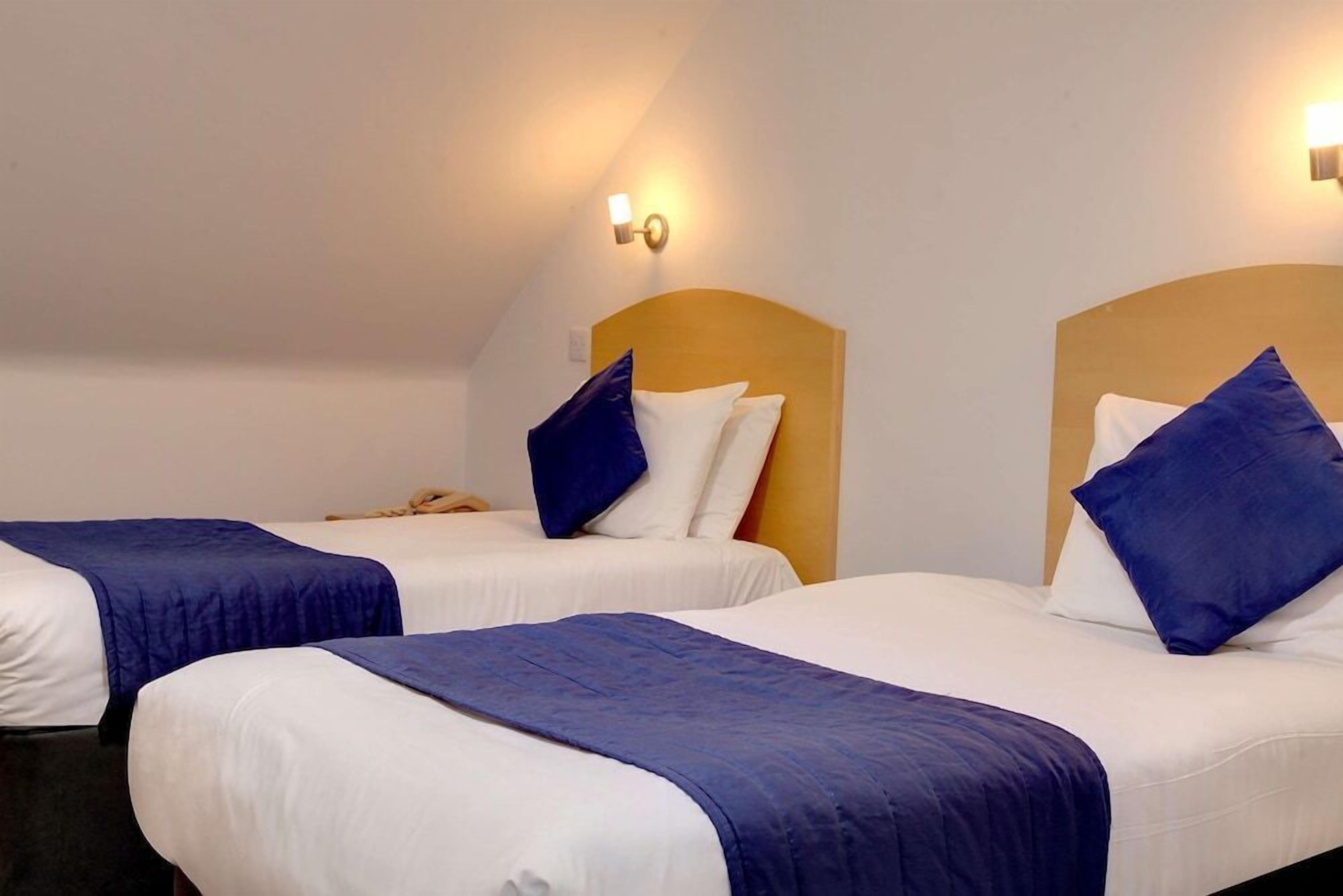 A hotel room with two single beds, each with a blue bedspread and white pillows. There are wall-mounted light fixtures on either side of the beds.