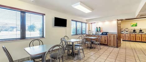 Un comedor amplio con varias mesas y sillas, una zona de cocina con armarios de madera y ventanas grandes con persianas.