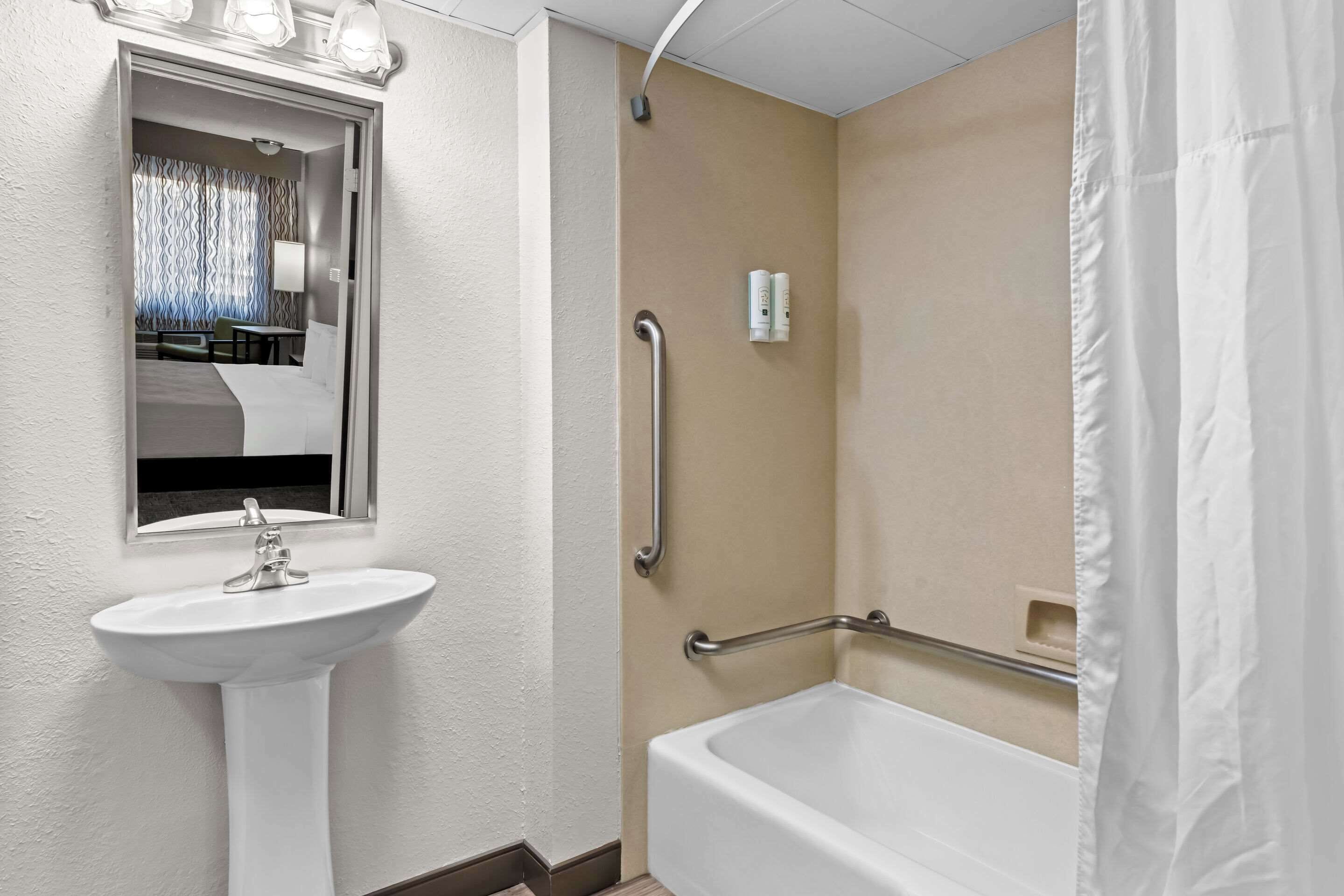 Une salle de bain avec un lavabo blanc, un miroir, une douche équipée d’une barre d’appui et une fenêtre avec des rideaux.