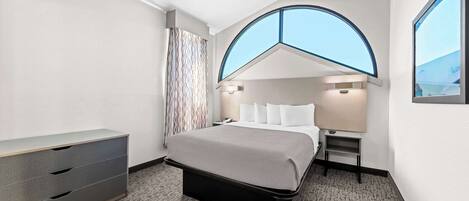 Una habitación de hotel moderna con una cama, un tocador, una mesita de noche y un ventanal grande.