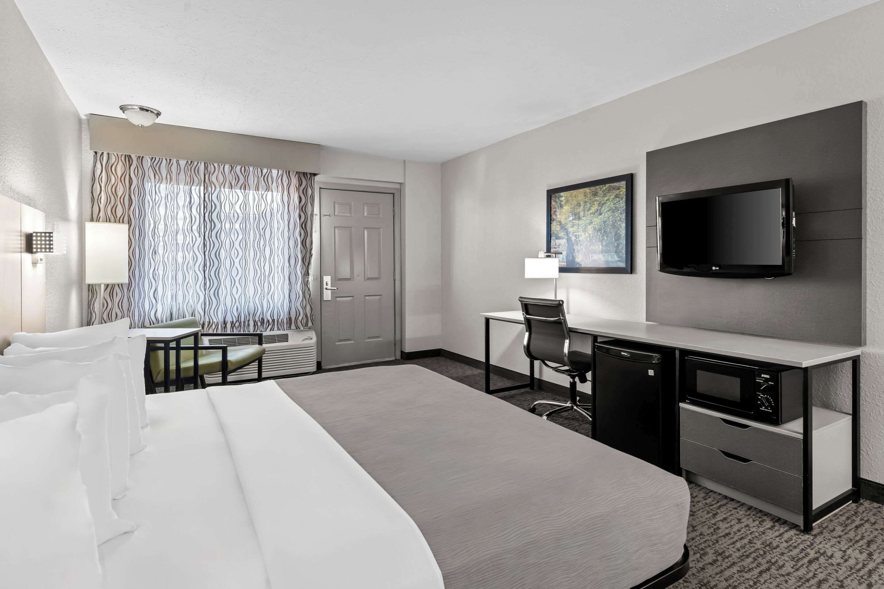 Habitación de hotel con una cama grande, un escritorio con computadora, un televisor de pantalla plana, una ventana con cortinas y una puerta.