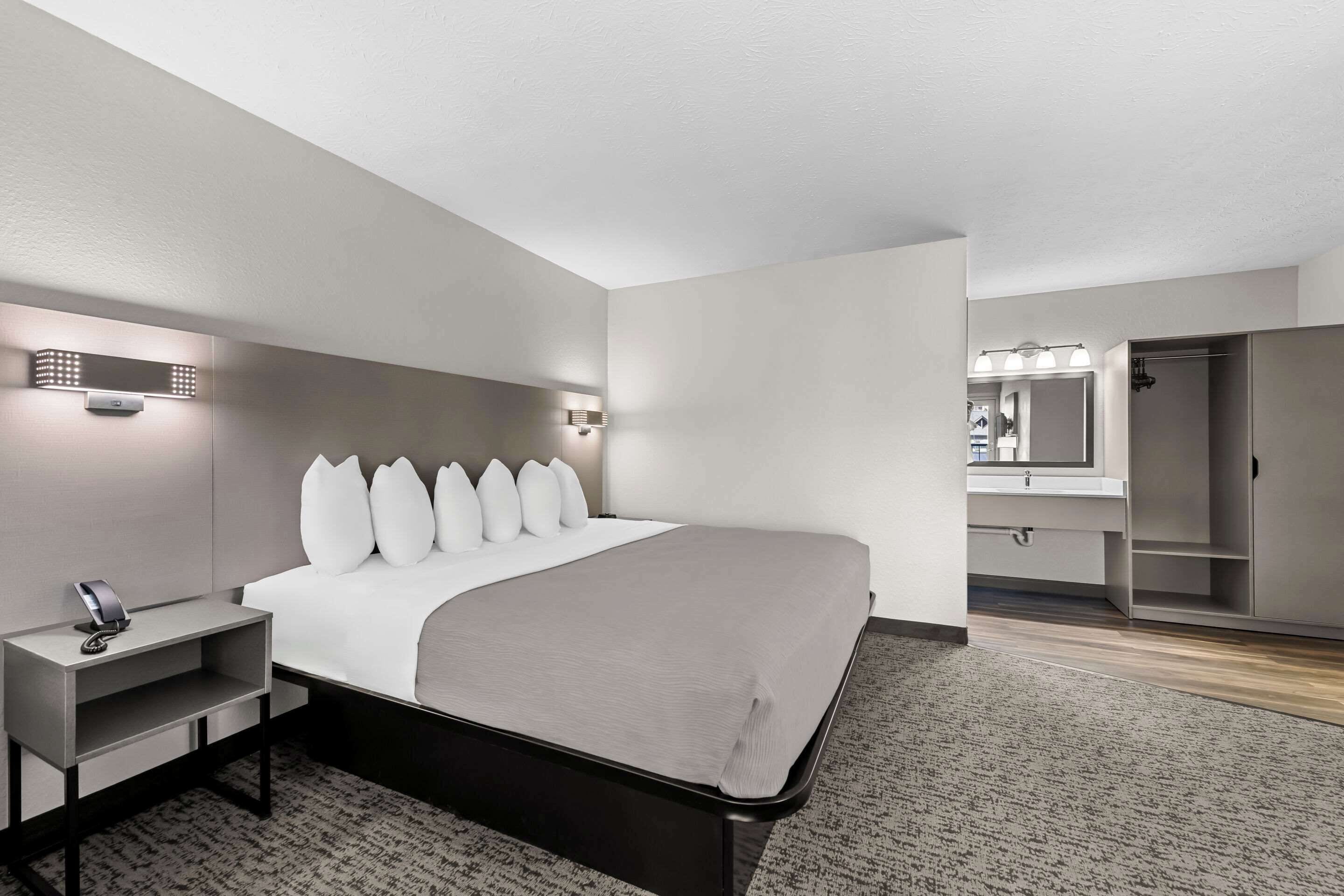A modern hotel room with a neatly made bed, a bedside table with a lamp, and a bathroom with a mirror and sink.