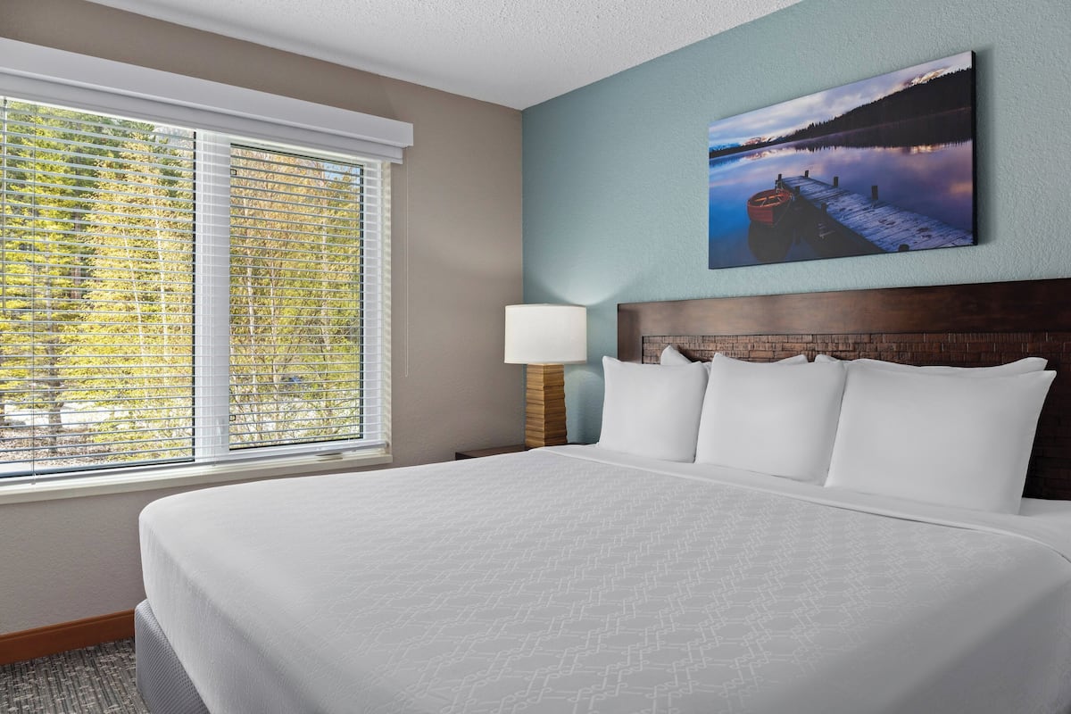 A hotel room with a large bed, a window with blinds, a bedside lamp, and a painting of a lake scene on the wall.
