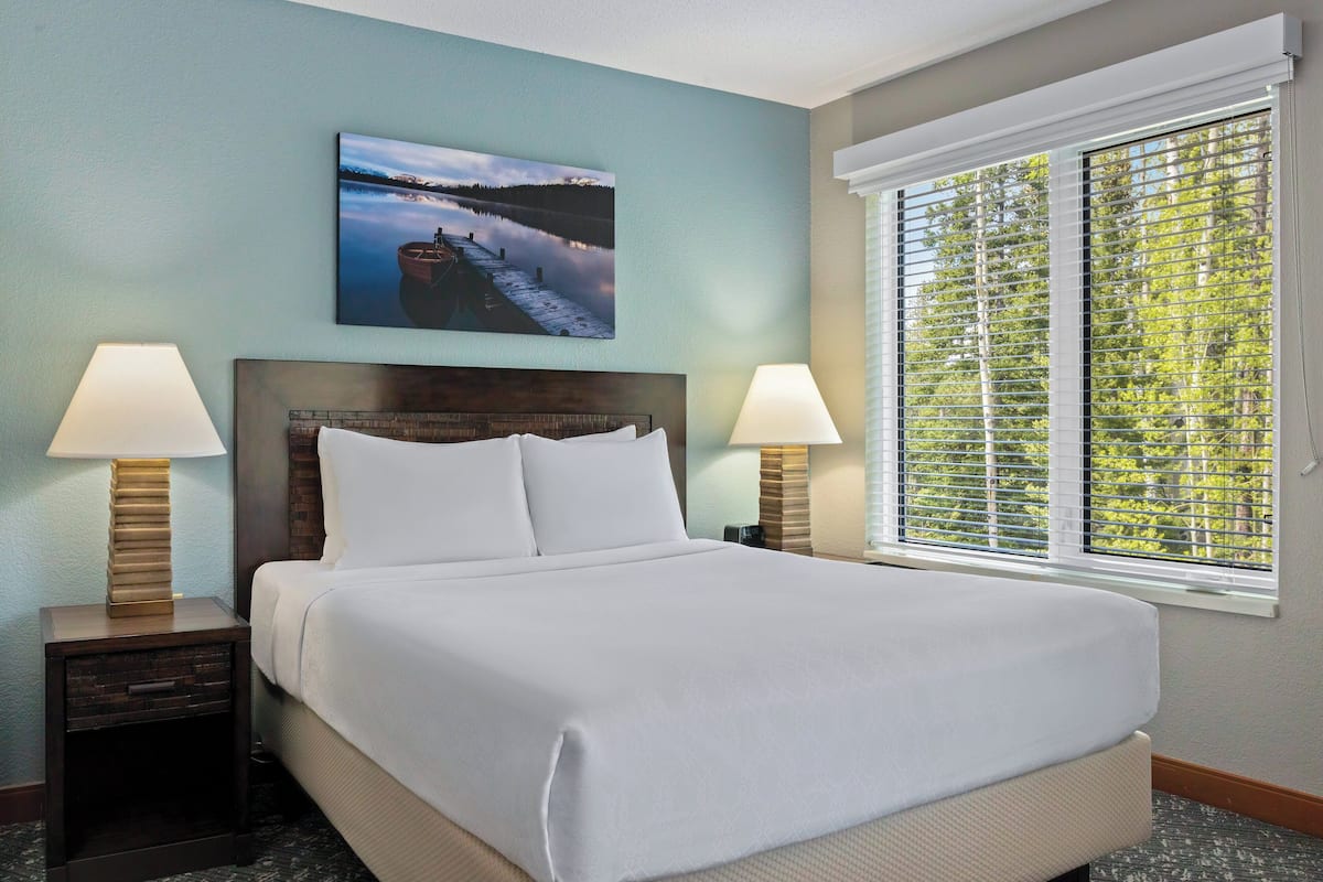A neatly made bed with white linens, two bedside lamps, a framed picture of a lake, and a window with blinds.