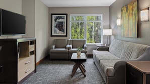A hotel room with a flat-screen TV, two armchairs, a sofa, a small table with a plant, a window with a view of trees, and a painting on the wall. - WorldMark Canmore - Banff (Canmore)