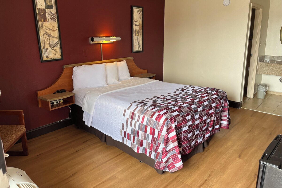 A hotel room with a wooden bed, a bedside table, a nightstand, a wall-mounted lamp, and a framed artwork above the bed.