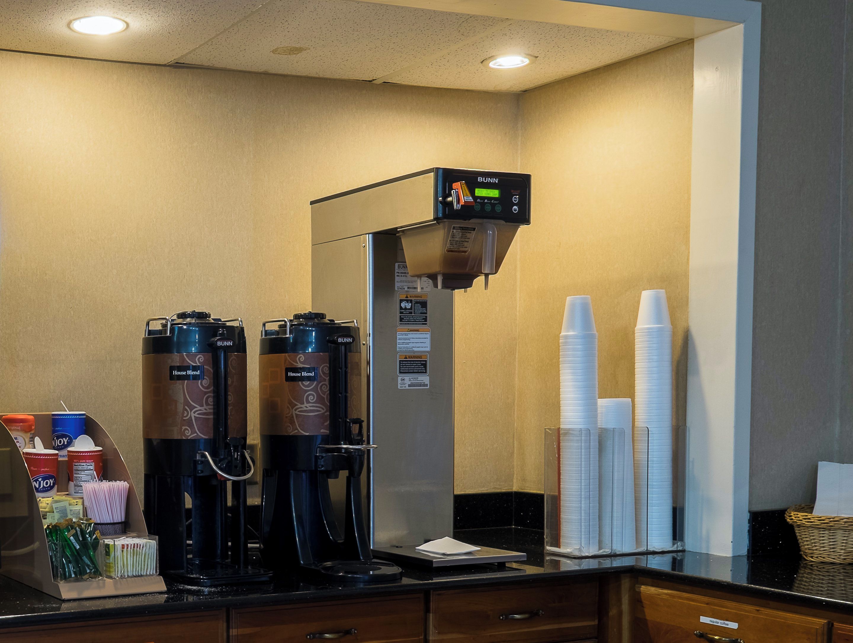 A coffee station with two Bunn coffee machines, a coffee dispenser, and a stack of disposable cups.