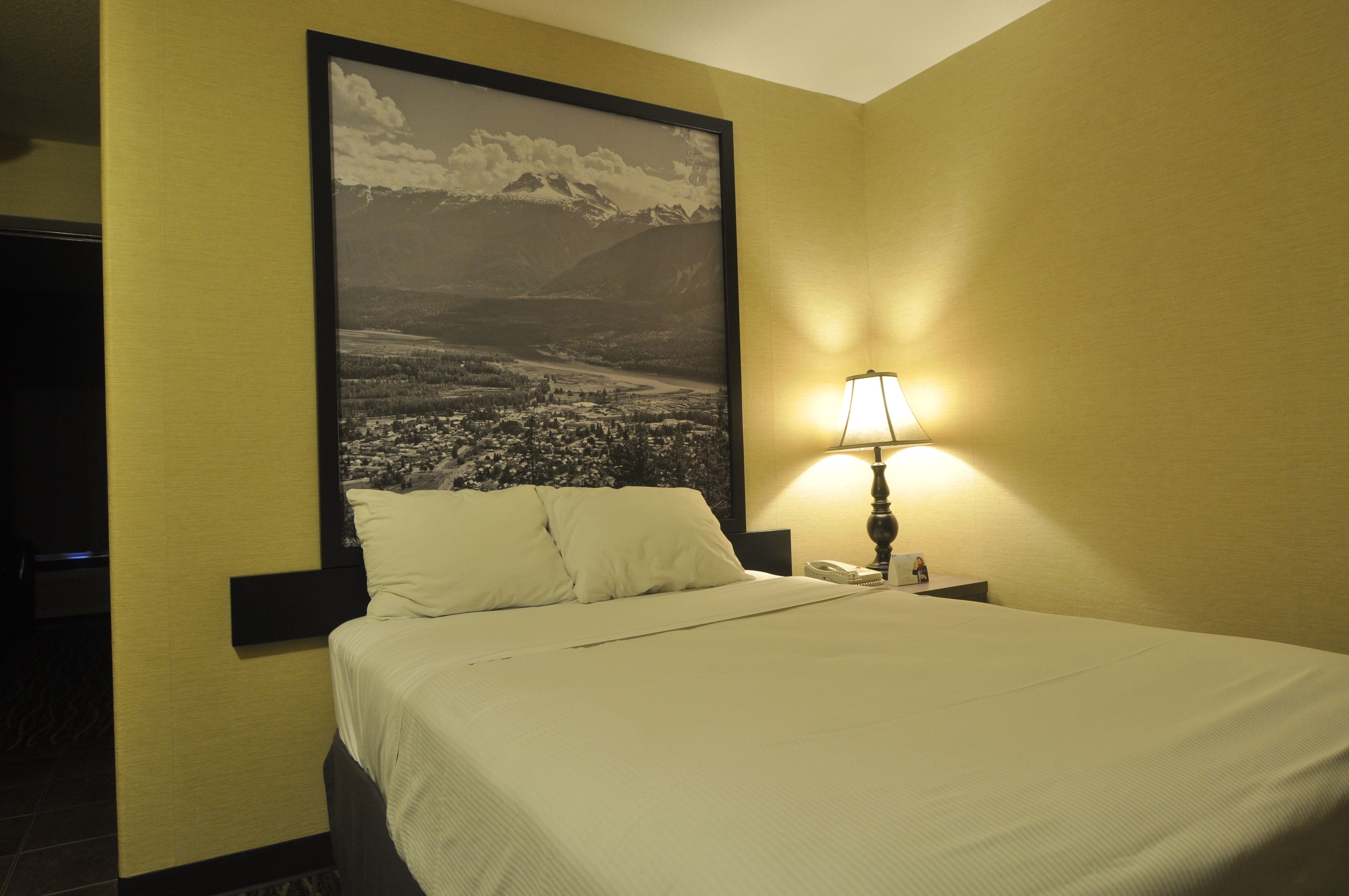Une chambre d’hôtel avec un lit, une table de chevet, une lampe et un grand tableau représentant un paysage en noir et blanc accroché au mur.