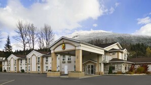 Un hotel con entrada cubierta, varias habitaciones y montañas al fondo.
