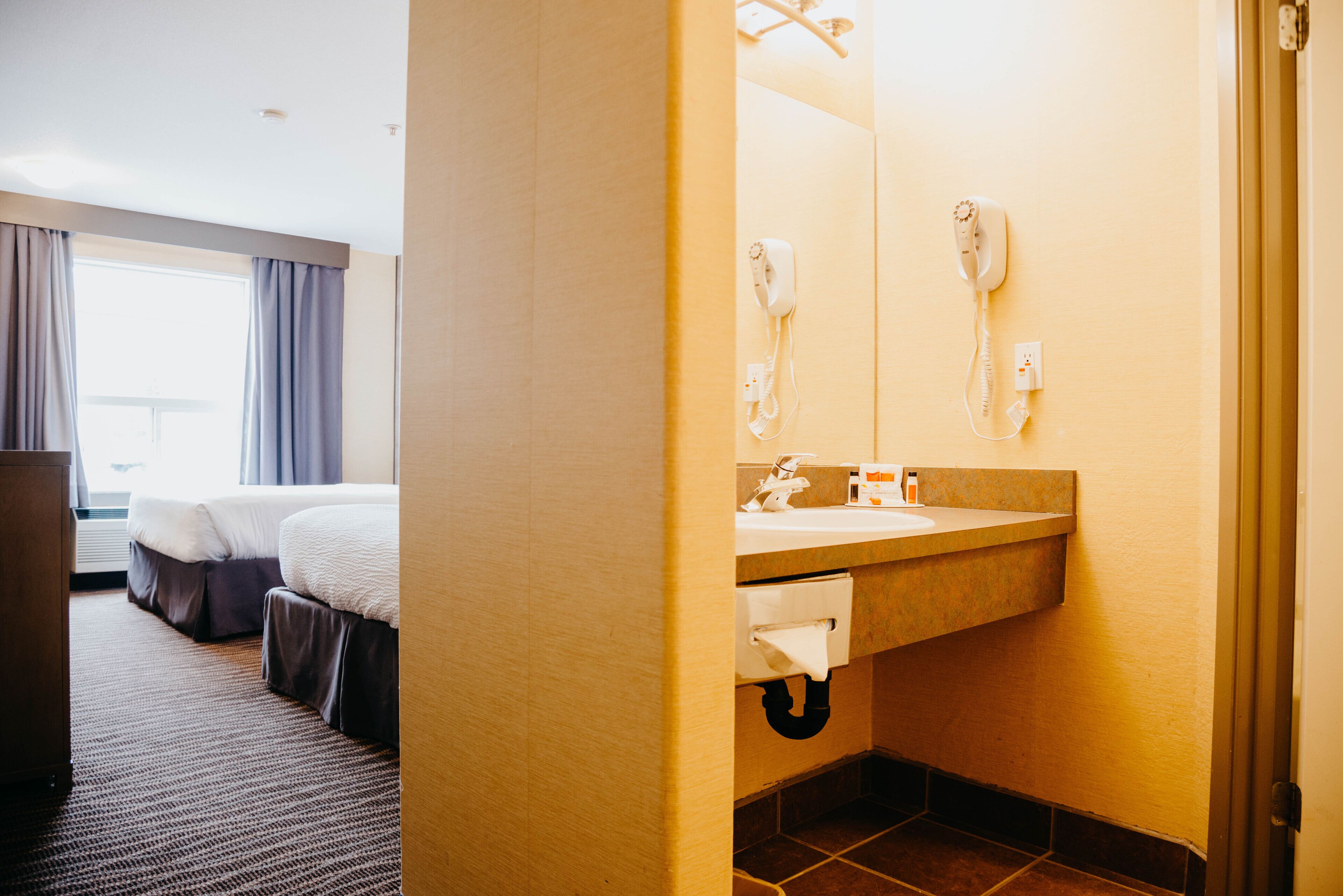 A hotel room with a bed, a bathroom with a sink and a hairdryer, and a window with curtains.
