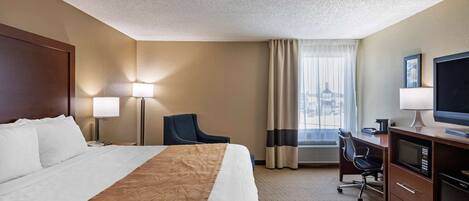 Habitación de hotel con una cama grande, un escritorio con computadora y televisión, una silla y una ventana con cortinas.