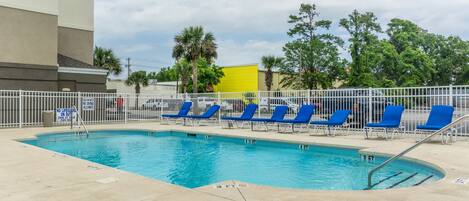 A swimming pool with blue lounge chairs, a white fence, and palm trees in the background.