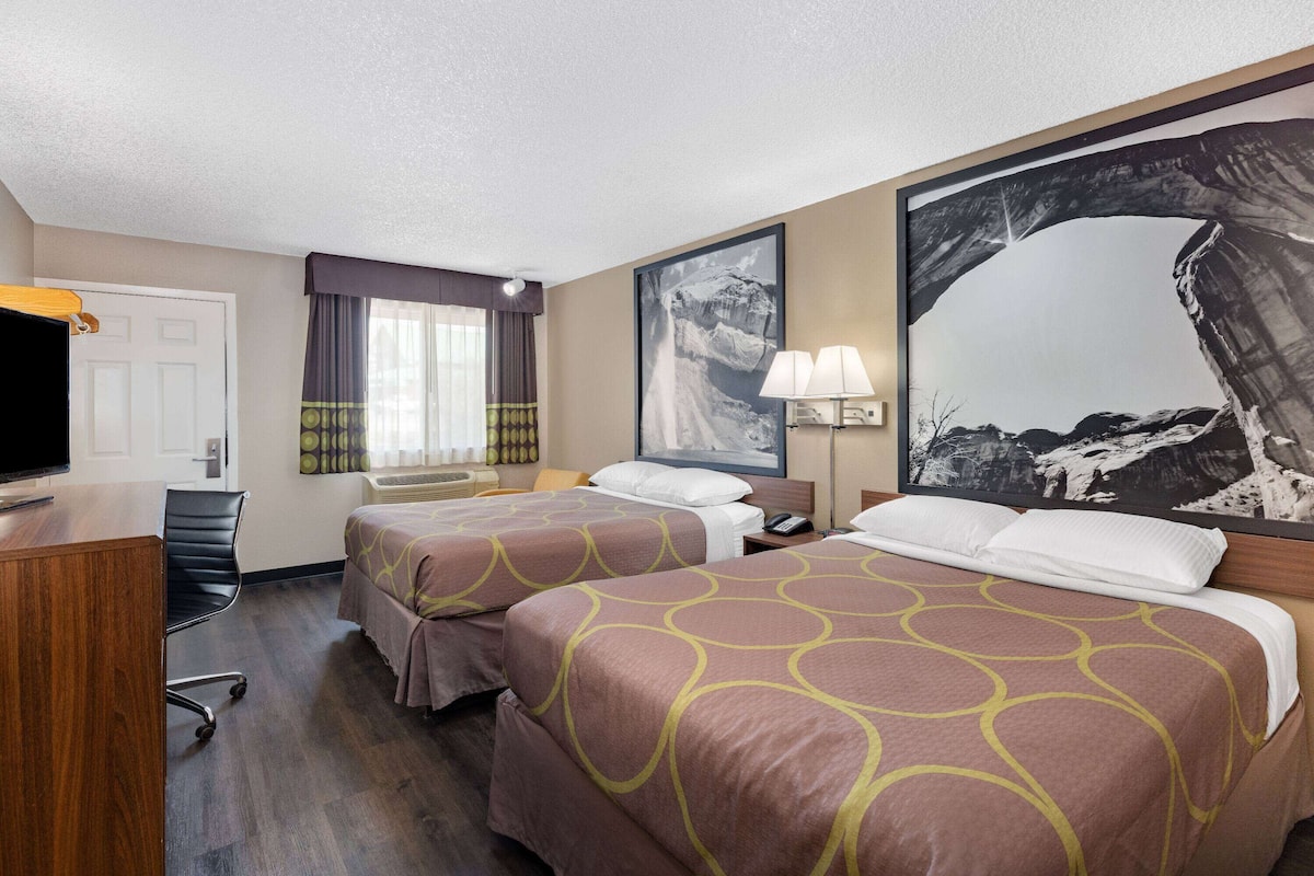 A hotel room with two beds, a desk, a chair, a television, a window with curtains, and a large framed picture on the wall.