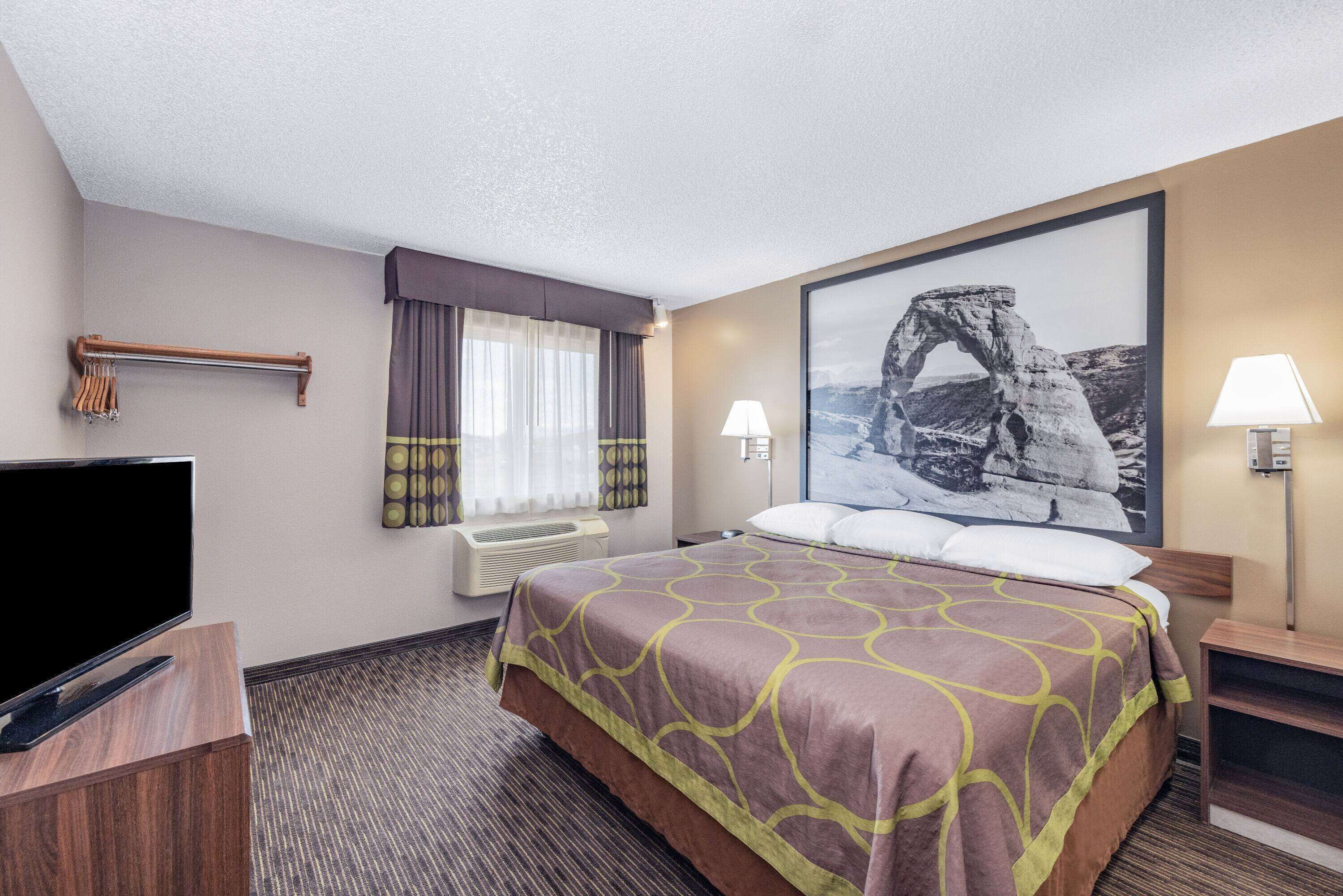 A hotel room with a bed, a flat-screen TV, a window with curtains, a wall-mounted lamp, and a framed picture of a rock formation.